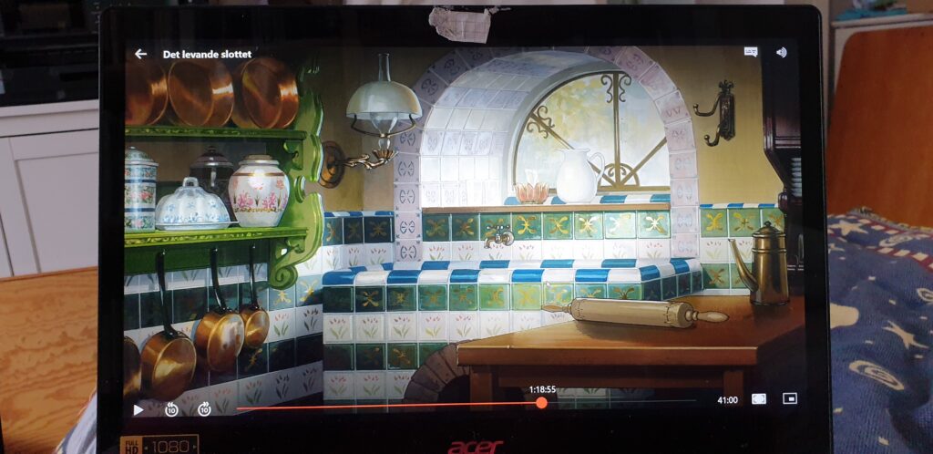 The wet corner now has a tiled sink, and the tiles are green or white with gold decorations. The green shelf has become more ornate and holds porcelain with elaborate flower motifs and copper cookware. On the other side a sturdy wooden workbench holds a rolling pin and a decorated coffee pot. An ornate kerosene lamp is mounted to the wall above the sink, and the window is enforced with gilded and decorated bars. 